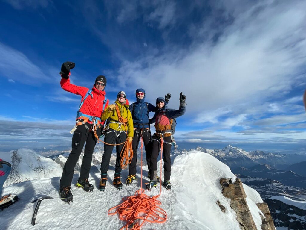 Vier Bergsteiger in Winterausrüstung stehen auf einem schneebedeckten Berggipfel, lächeln und recken jubelnd die Arme in die Höhe, umgeben von Seilen und Klettergurten nach einer erfolgreichen Besteigung der Dom- und Dufourspitze, im Hintergrund erheben sich malerische Berge vor einem blauen Himmel.