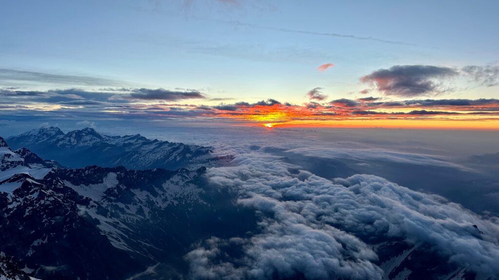Ein spektakulärer Sonnenaufgang über schneebedeckten Bergen, darunter der majestätische Dom und die Dufourspitze, während Wolken die Täler darunter ausfüllen und ein leuchtend orangefarbenes Licht den Himmel in der Ferne erhellt.