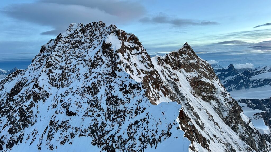 Schneebedeckte Berggipfel unter einem teilweise bewölkten Himmel, mit schroffen, felsigen Hängen und fernen Bergen im Hintergrund – ein inspirierender Anblick, den man oft auf einer geführten Tour zur Dom- und Dufourspitze erleben kann.