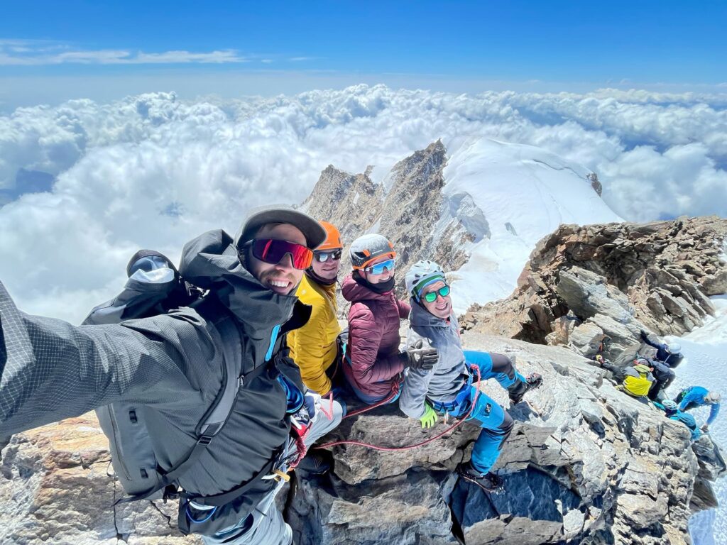 Vier Bergsteiger in Winterausrüstung lächeln für ein Selfie auf einem verschneiten Berggipfel entlang der berühmten Spaghetti-Tour am Monte Rosa, während unter ihnen felsiges Gelände und Wolken unter einem strahlend blauen Himmel zu sehen sind.