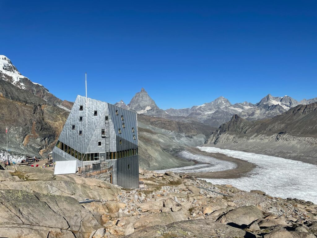 Ein modernes, kantiges Metallgebäude steht auf felsigem Gelände mit Blick auf einen Gletscher, im Hintergrund schneebedeckte Berge und ein strahlend blauer Himmel – ein beeindruckender Zwischenstopp auf der legendären Spaghetti-Tour Monte Rosa.