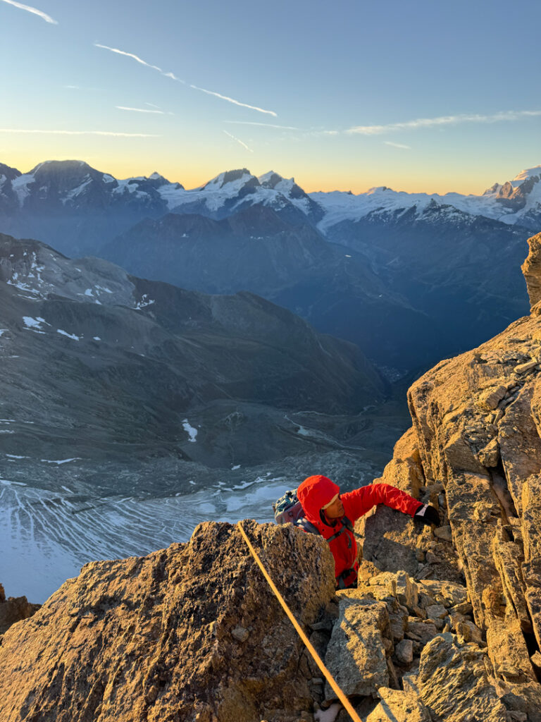 Ein Bergsteiger in roter Ausrüstung erklimmt bei Sonnenaufgang einen felsigen Berg – möglicherweise auf einer geführten Route vom Obergabelhorn zum Zinalrothorn –, während unter dem klaren Himmel Schneefelder, ferne Gipfel und ein tiefes Tal zu sehen sind.
