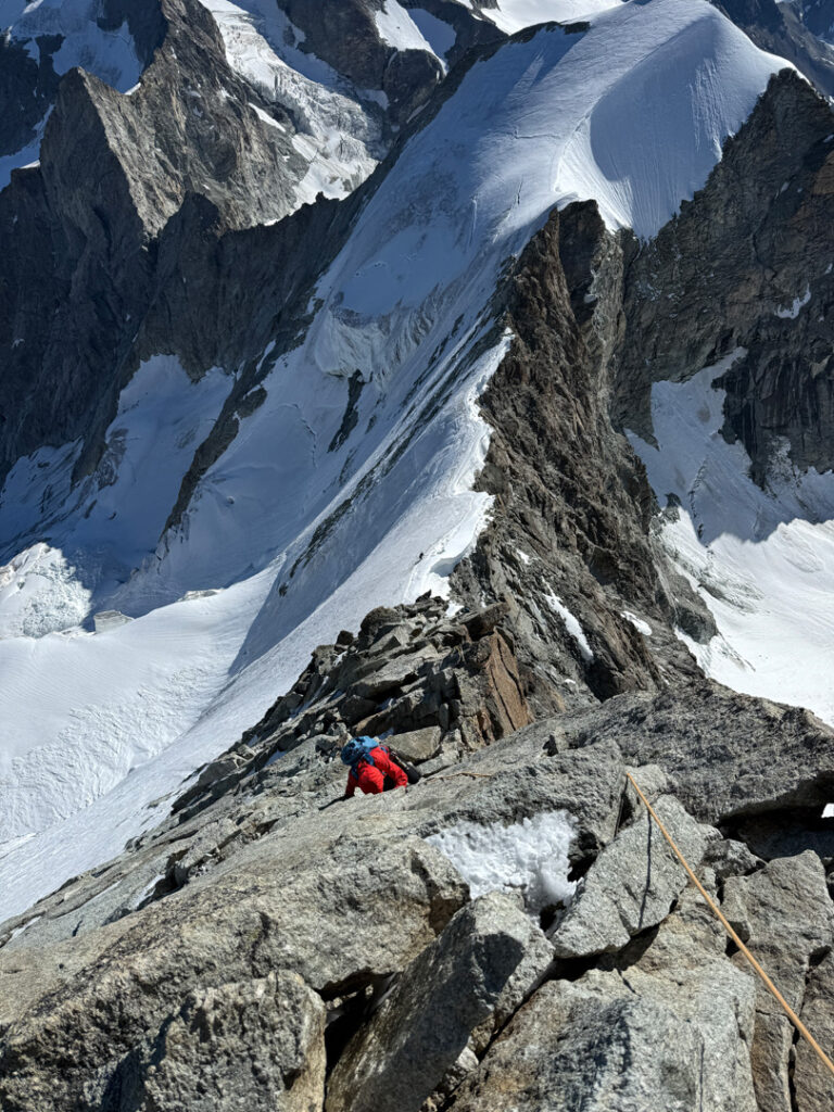 Ein Bergsteiger in roter Kleidung erklimmt einen felsigen, schneebedeckten Berggrat in der Nähe des Obergabelhorns und des Zinalrothorns und überwindet dabei steile Hänge und schroffe Gipfel bei strahlendem Tageslicht.