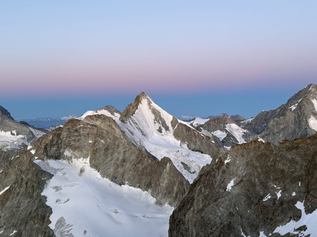 Ein Blick auf schroffe, schneebedeckte Alpengipfel wie das Obergabelhorn und das Zinalrothorn, deren majestätische Gipfel sich vor einem Abendhimmel erheben, der am Horizont von tiefem Blau in ein sanftes Rosa übergeht. Gletscher füllen die Täler zwischen den schroffen, dunklen Felswänden und schaffen eine atemberaubende Kulisse, die sich ideal für ein geführtes Abenteuer eignet.