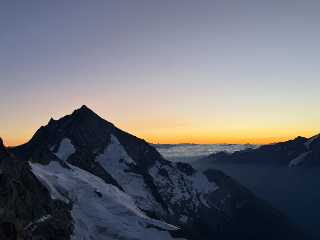 Das schneebedeckte Weisshorn zeichnet sich bei Sonnenaufgang als Bergspitze ab, über einer Wolkendecke und fernen Gebirgszügen erstreckt sich ein leuchtend orange-gelber Himmel.