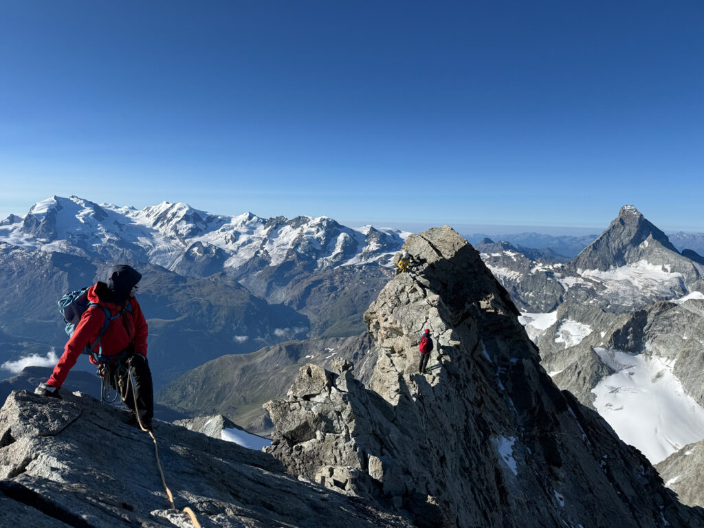 Eine Gruppe von Bergsteigern in roten Jacken erklimmt mit Seilen einen felsigen, verschneiten Berggrat im Rahmen einer anspruchsvollen geführten Tour vom Obergabelhorn zum Zinalrothorn. Im Hintergrund erstrecken sich schneebedeckte Gipfel und ein strahlend blauer Himmel.