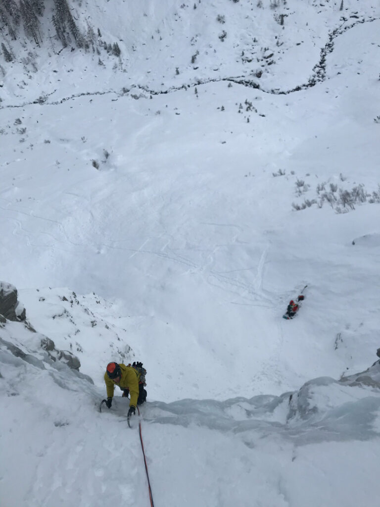 Ein Kletterer in einer gelben Jacke erklimmt eine vereiste, schneebedeckte Felswand, gesichert durch ein rotes Seil; weiter unten auf dem verschneiten Boden sind eine weitere Person und Eiskletterausrüstung zu sehen.