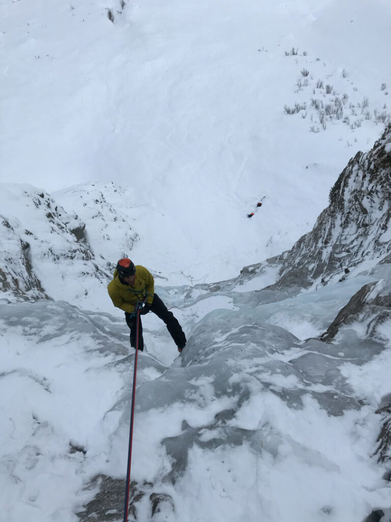 Eine Person in einer gelben Jacke klettert mit einem Seil einen verschneiten, vereisten Hang hinauf, umgeben von steilen, schneebedeckten Klippen und Felsen, und demonstriert dabei die anspruchsvollen Techniken des Eiskletterns.