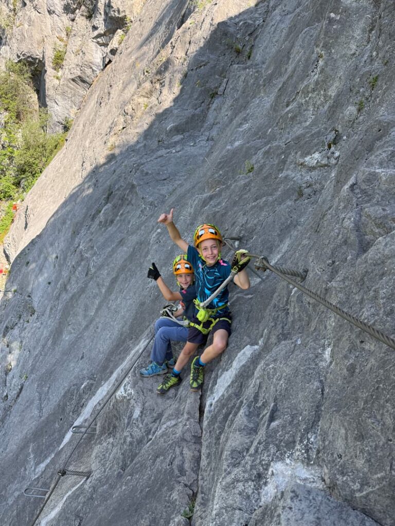 Zwei Kinder, die Helme und Klettergurte tragen, sitzen auf einer steilen Felswand, halten sich an einem Sicherheitsseil fest und zeigen den Daumen nach oben, während sie einen der geführten Klettersteige im Freien erkunden.