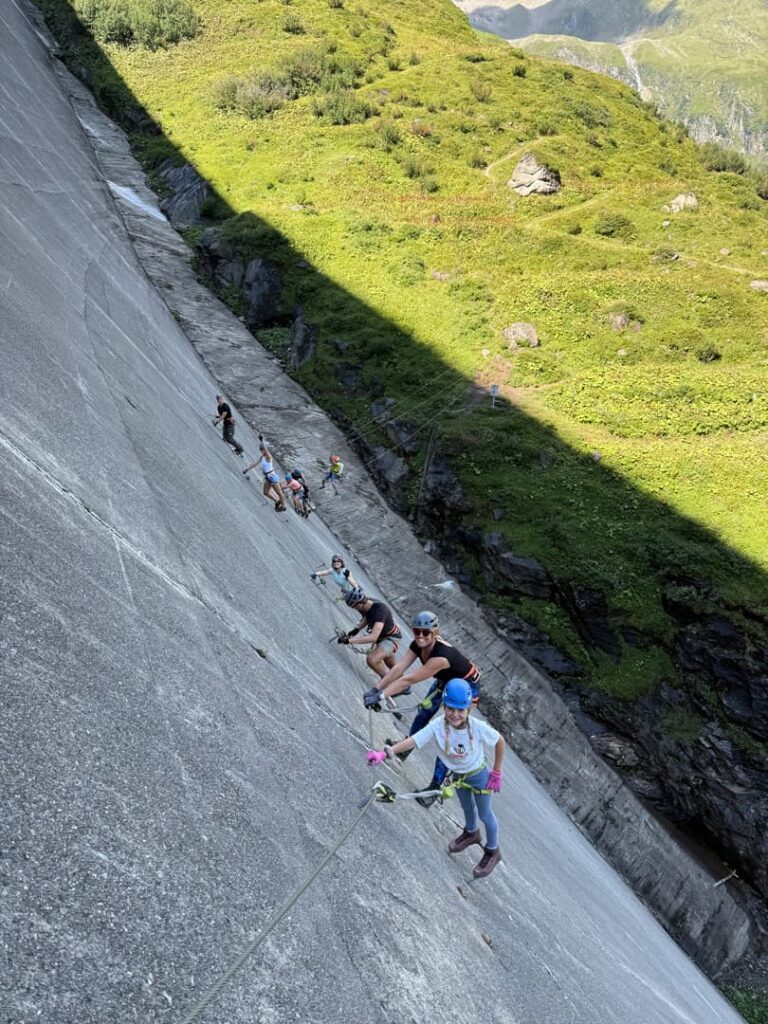 Eine Gruppe von Menschen in Kletterausrüstung erklimmt unter strahlendem Sonnenschein eine steile Betonwand neben einem grasbewachsenen Berghang und nutzt dabei ein Sicherheitsseil als Halt – als Teil eines geführten Klettersteigs.