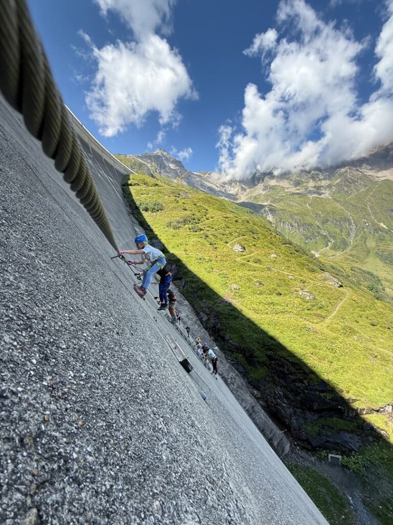 Menschen mit Helmen und Klettergurten erklimmen auf geführten Klettersteigen mithilfe von Seilen und Metallstufen einen steilen Betonhang, vor dem sich grüne Berge und ein blauer Himmel mit Wolken abzeichnen.