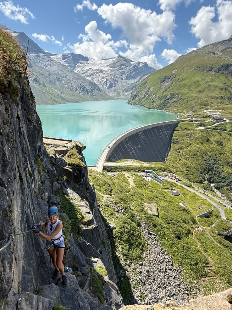 Ein Kletterer mit Helm und Klettergurt erklimmt eine felsige Klippe über einem üppig grünen Tal und nimmt an einer geführten Klettersteigtour teil, während sich unter ihm ein großer Staudamm und ein türkisfarbener Stausee erstrecken und im Hintergrund unter einem teilweise bewölkten Himmel schneebedeckte Berge zu sehen sind.