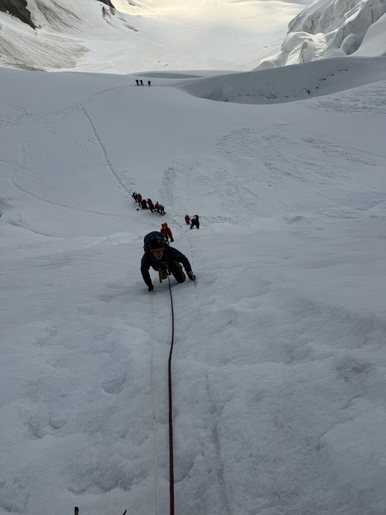 Ein Bergsteiger erklimmt auf dieser geführten Expedition mithilfe eines Seils einen steilen, verschneiten Hang, gefolgt von anderen, die hinter ihm in einer Reihe hergehen. Im Hintergrund sind große, schneebedeckte Berge – wie der Eiger, der Mönch und die Jungfrau – sowie Spuren zu sehen.
