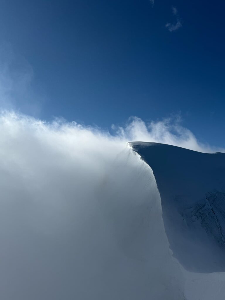 Ein schneebedeckter Berggipfel mit einem schroffen Grat, teilweise in Nebel gehüllt, unter strahlend blauem Himmel. Das Sonnenlicht lässt den Rand des verschneiten Hangs erstrahlen und erinnert an die dramatischen Ausblicke, die man auf einer geführten Alpintour entlang des Eigers und des Mönchs genießen kann.