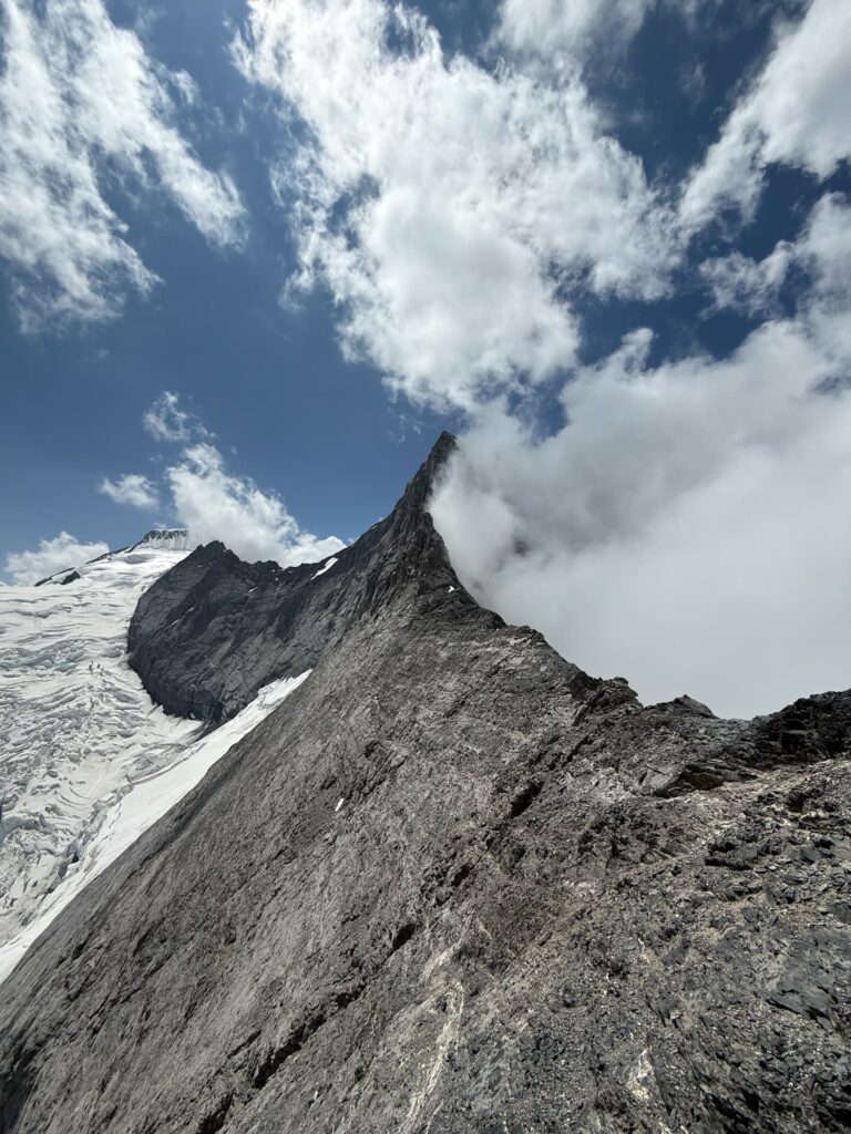 Ein steiler, felsiger Bergrücken mit Schneeflecken, teilweise von Wolken verdeckt, unter einem strahlend blauen Himmel mit vereinzelten Wolken – die legendären Gipfel Eiger, Mönch und Jungfrau ragen majestätisch entlang dieser klassischen Alpinroute empor, die oft im Rahmen von geführten Touren erkundet wird.