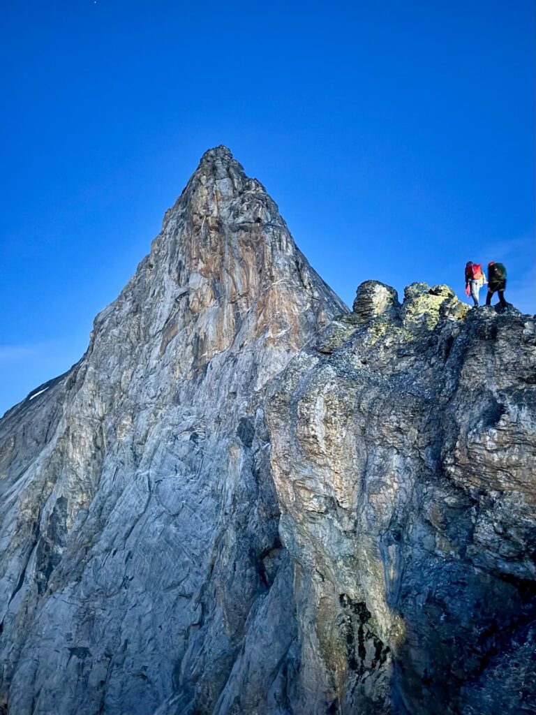Zwei Wanderer mit Rucksäcken stehen auf einem felsigen Berggrat unter strahlend blauem Himmel, während sich neben ihnen die steilen, zerklüfteten Gipfel von Eiger, Mönch und Jungfrau erheben.