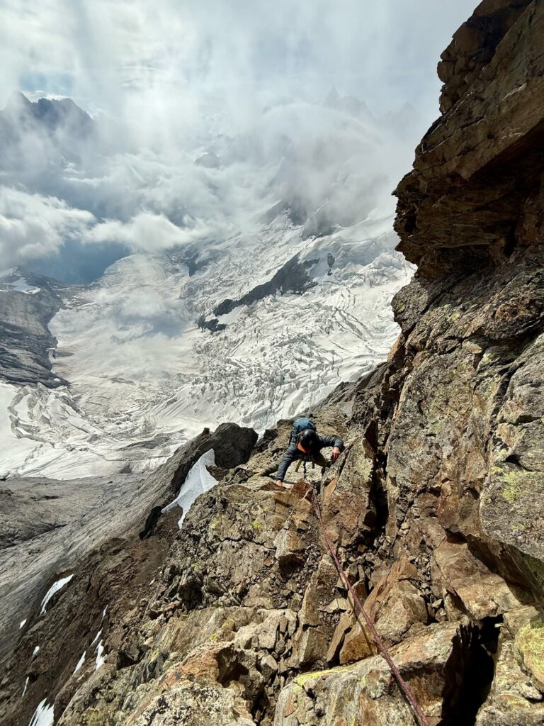 Ein Kletterer erklimmt eine steile Felswand, sicher angeseilt im Rahmen einer geführten Tour, während sich im Hintergrund die legendären, schneebedeckten Gipfel von Eiger, Mönch und Jungfrau majestätisch aus den vorbeiziehenden Wolken erheben.