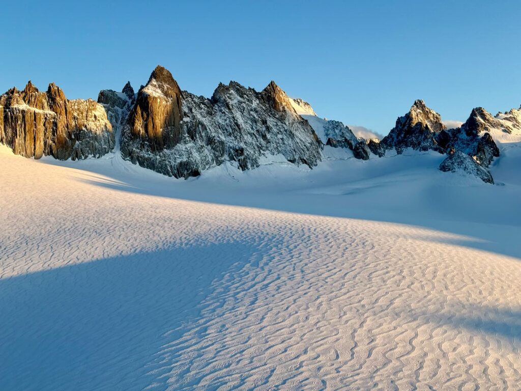 Schneebedeckte Berge unter strahlend blauem Himmel, wobei das Sonnenlicht Schatten auf den welligen Schnee im Vordergrund wirft – eine Szene, die an eine geführte Mont-Blanc-Expedition erinnert.