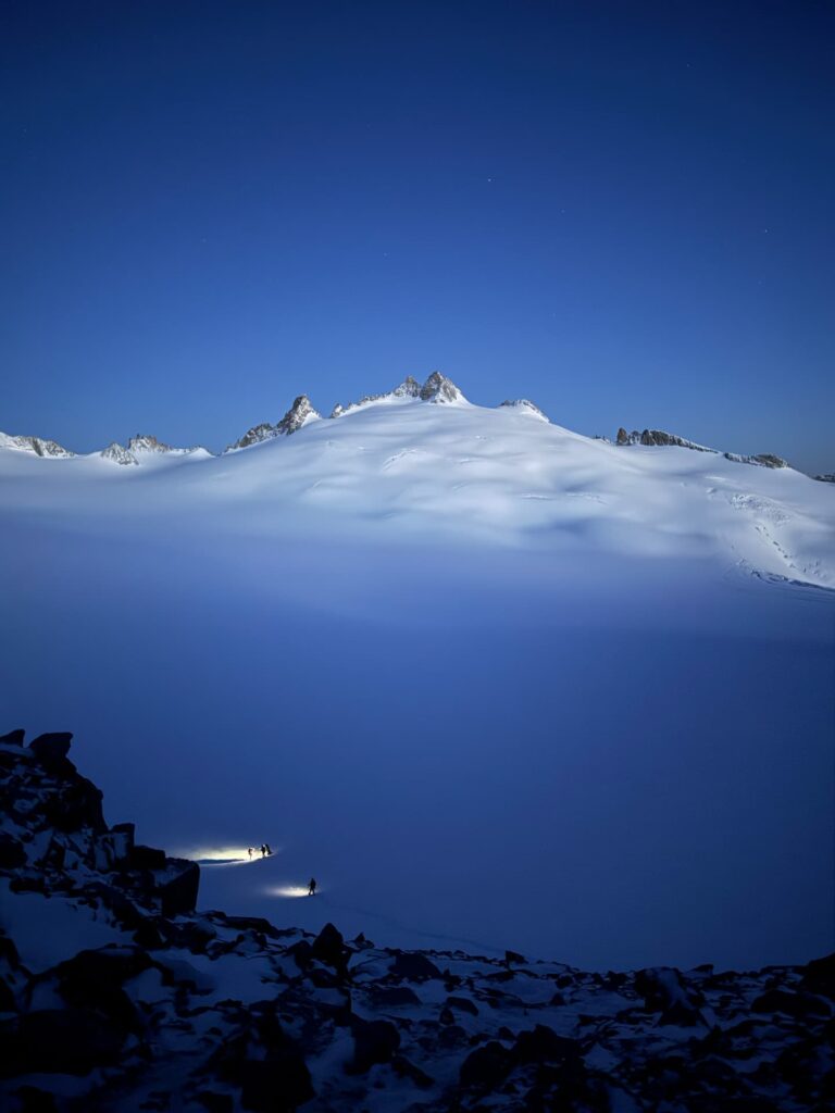 Zwei kleine Gestalten mit Stirnlampen durchqueren in der Abenddämmerung unter einem tiefblauen Himmel eine weite, verschneite Fläche und bewegen sich auf ferne Berggipfel zu – vielleicht auf einer geführten Mont-Blanc-Expedition. In der unteren linken Ecke ist felsiges Gelände zu erkennen.
