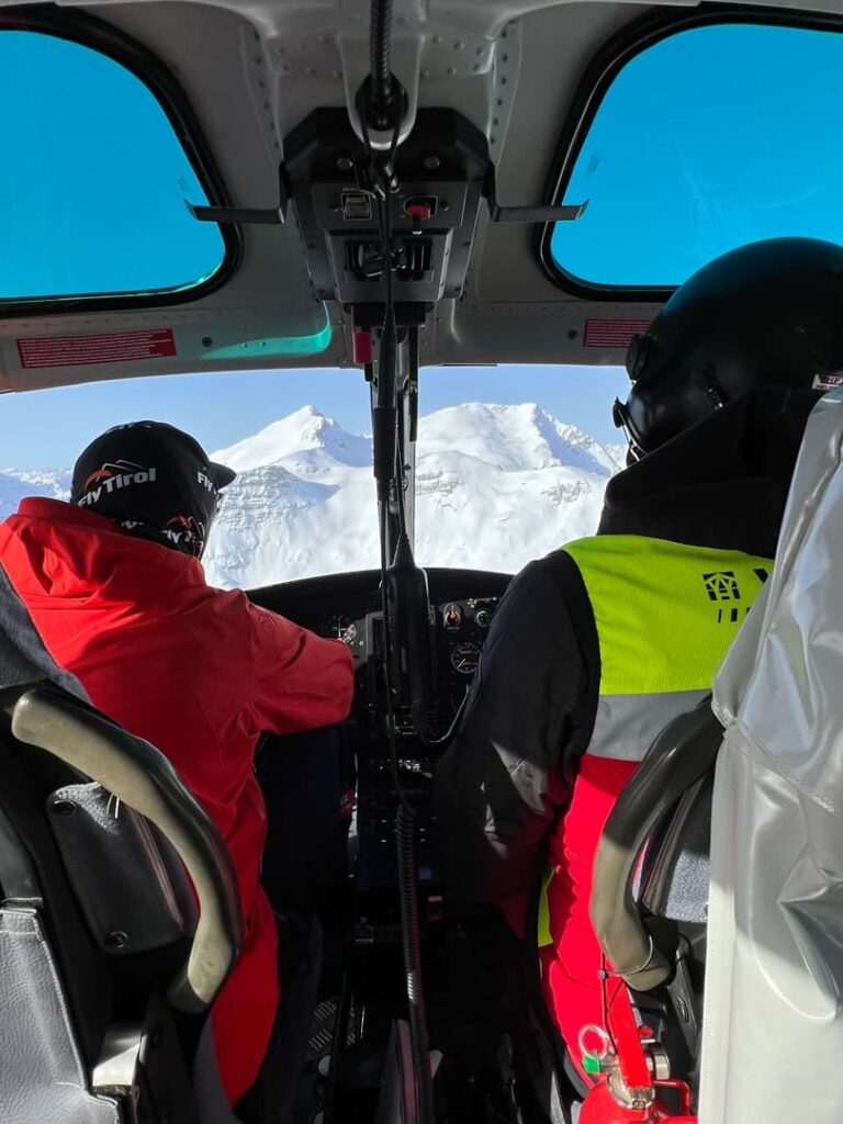 Zwei Personen in Winterkleidung sitzen im Cockpit eines Hubschraubers und fliegen unter strahlend blauem Himmel auf schneebedeckte Berge zu – bereit für spannende Abenteuer bei „Freeride Touren Alpen“.