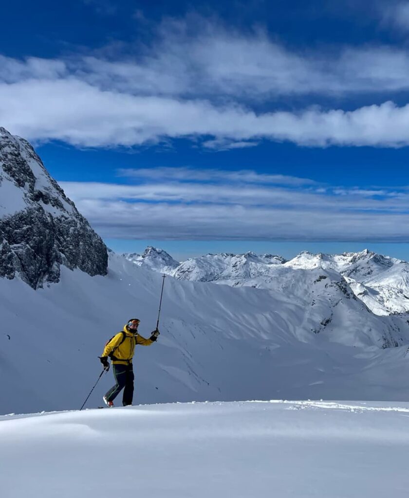 Ein Skifahrer in einer gelben Jacke steht auf einem verschneiten Berghang, bereit für geführte Skitouren, und hält Skistöcke in der Hand; im Hintergrund sind schneebedeckte Gipfel und ein blauer Himmel mit Wolken zu sehen.