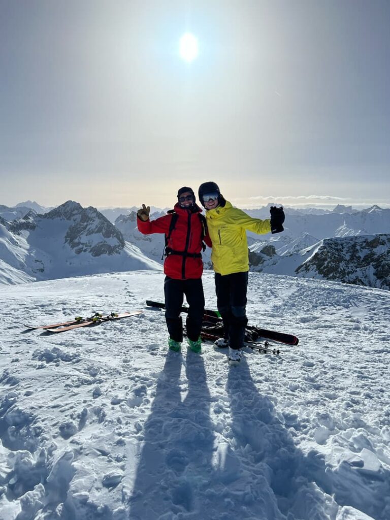 Zwei Personen in Skikleidung stehen auf einem verschneiten Berggipfel, lächeln und posieren mit erhobenen Armen, nachdem sie eine der aufregenden Freeride-Touren in den Alpen absolviert haben. Hinter ihnen liegen die Skier auf dem Boden, während die Sonne strahlend vom Himmel scheint und lange Schatten auf den Schnee wirft. Im Hintergrund erstrecken sich die Berggipfel und fangen die atemberaubende Kulisse ihres alpinen Abenteuers ein.