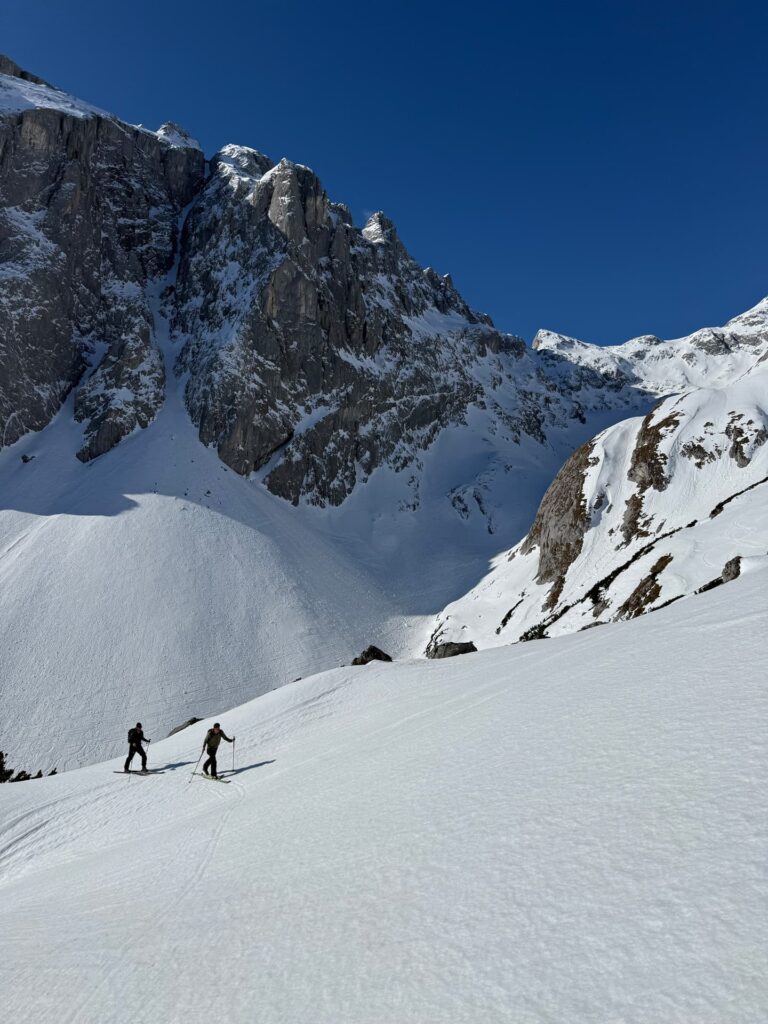 Zwei Menschen wandern einen verschneiten Berghang hinauf, umgeben von steilen, schneebedeckten Felsen und unter einem strahlend blauen Himmel – eine klassische Szene aus den Abenteuern von „Freeride Touren Alpen“.