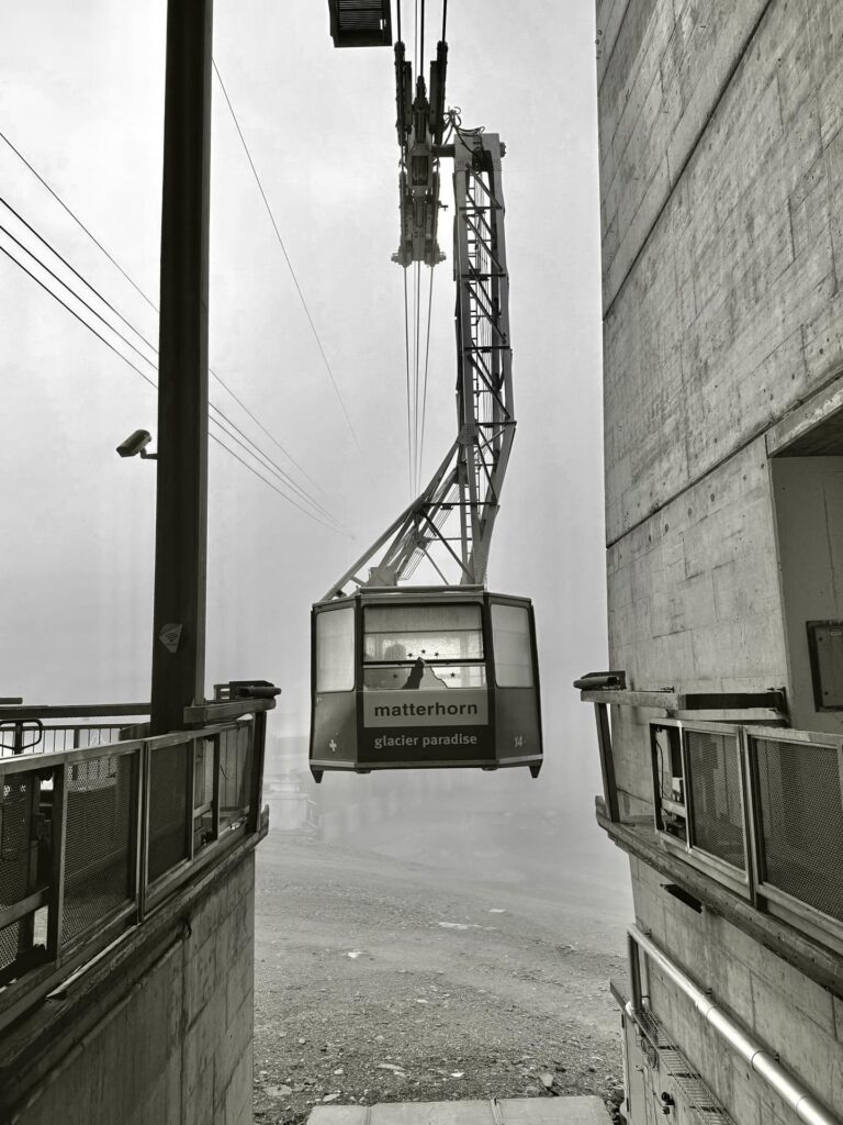 Eine Seilbahn mit der Aufschrift „Matterhorn glacier paradise“ schwebt an einem nebligen Tag zwischen zwei Betonkonstruktionen in der Luft, als würde sie still auf das nächste „Matterhorn geführt“-Abenteuer durch den Nebel warten.
