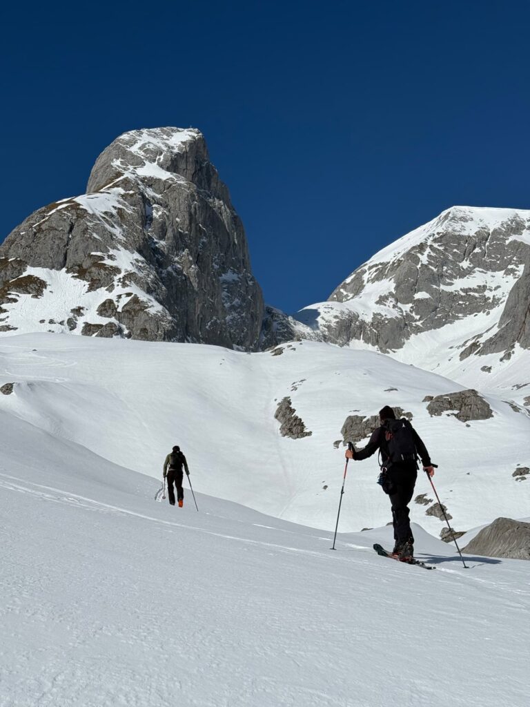 Zwei Personen in Winterkleidung steigen mit Skistöcken an einem verschneiten Berghang hinauf. Diese Szene verkörpert den Geist der geführten Skidurchquerungen in den Alpen, während sich im Hintergrund felsige, schneebedeckte Gipfel steil vor einem strahlend blauen Himmel erheben.