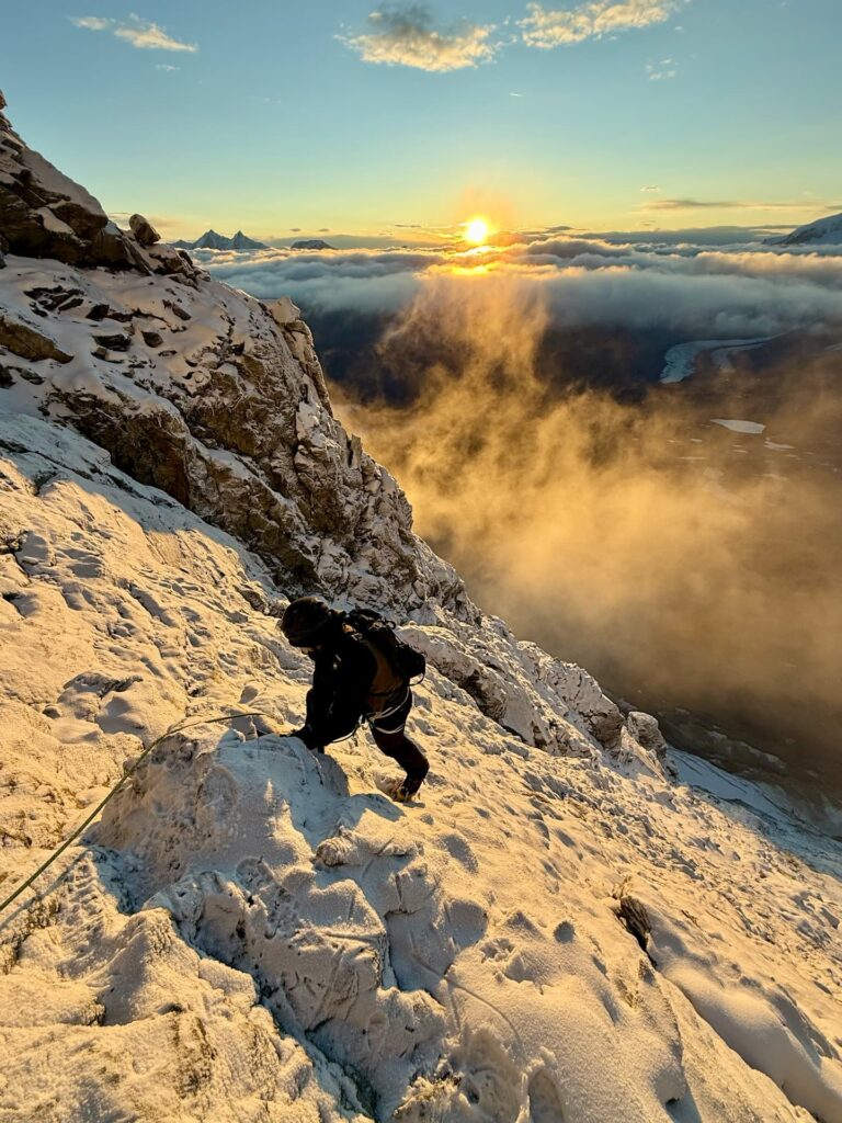 Ein Bergsteiger erklimmt bei Sonnenaufgang einen verschneiten Berghang, während goldenes Sonnenlicht die Wolken und schroffen Gipfel im Hintergrund beleuchtet – eine beeindruckende Szene, die an ein klassisches „Matterhorn geführt“-Abenteuer erinnert.