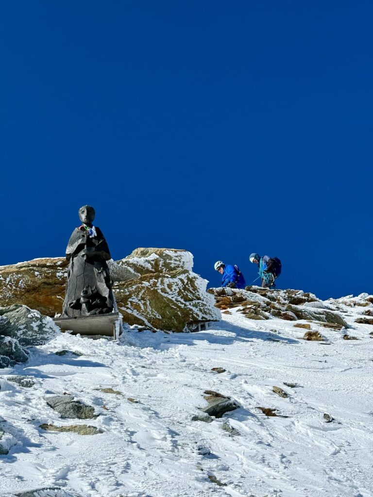 Auf einem verschneiten Berghang steht eine Statue, während zwei Bergsteiger mit Helmen und Ausrüstung unter strahlend blauem Himmel in der Nähe aufsteigen – ein Bild, das den Geist eines „Matterhorn geführt“-Abenteuers einfängt.