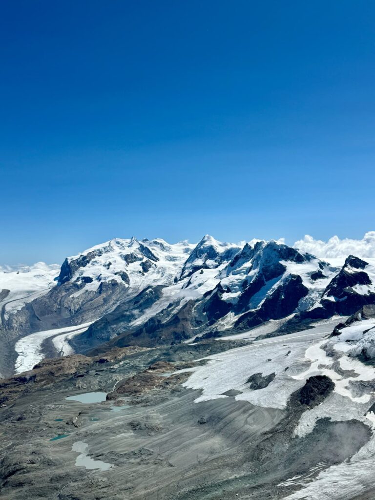 Schneebedeckte Berge unter strahlend blauem Himmel, im Vordergrund Gletscher, felsiges Gelände und kleine Seen – eine ikonische Landschaft, die an eine geführte Matterhorn-Tour erinnert.
