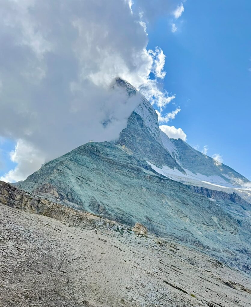 Ein felsiger Berggipfel mit Schneeflecken ist vor einem strahlend blauen Himmel teilweise von Wolken verhüllt. Im Vordergrund erstreckt sich ein felsiger, karger Hang – ein atemberaubender Anblick, den man oft bei einer geführten Besteigung des Matterhorns erleben kann.
