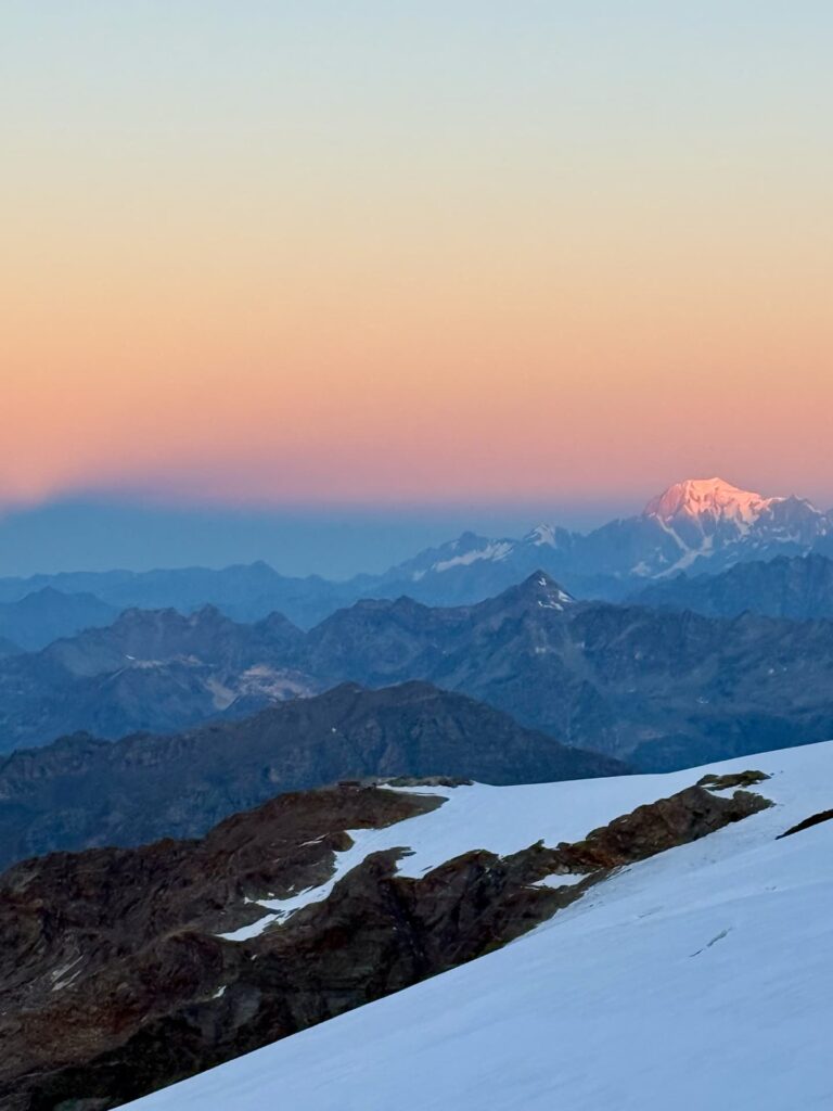 Sonnenuntergang über schneebedeckten Berggipfeln während der Spaghetti-Tour Monte Rosa, mit einem pastellorangen- und rosafarbenen Himmel. Ein entfernter Gipfel wird vom Sonnenlicht beleuchtet, während die Berge im Vordergrund im Schatten liegen. Im Vordergrund sind Schnee und felsige Grate zu sehen.