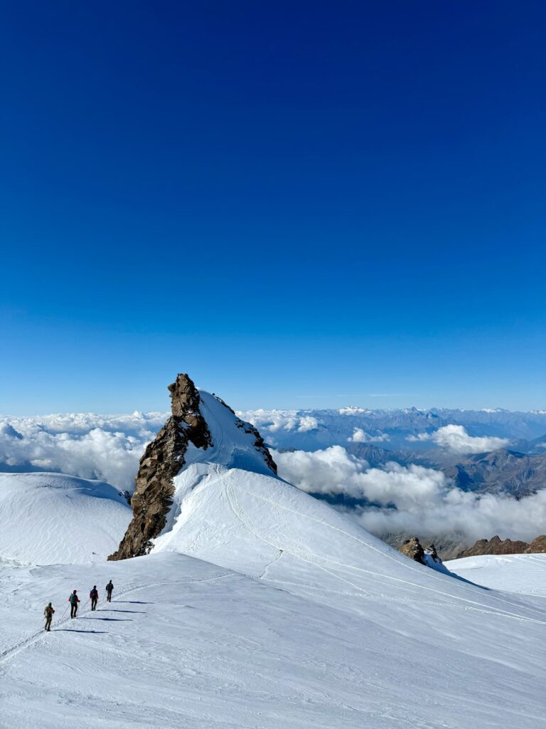 Vier Menschen wandern unter strahlend blauem Himmel über einen verschneiten Bergrücken in Richtung eines felsigen Gipfels, im Hintergrund sind ferne Berge und Wolken zu sehen – sie begeben sich auf die berühmte Spaghetti-Tour am Monte Rosa.