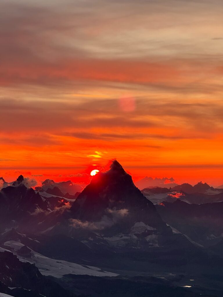 Die Sonne geht hinter einem schroffen Berggipfel entlang der berühmten Spaghetti-Tour am Monte Rosa unter, wirft dramatische Schatten und taucht den Himmel in leuchtende Orange- und Rottöne.