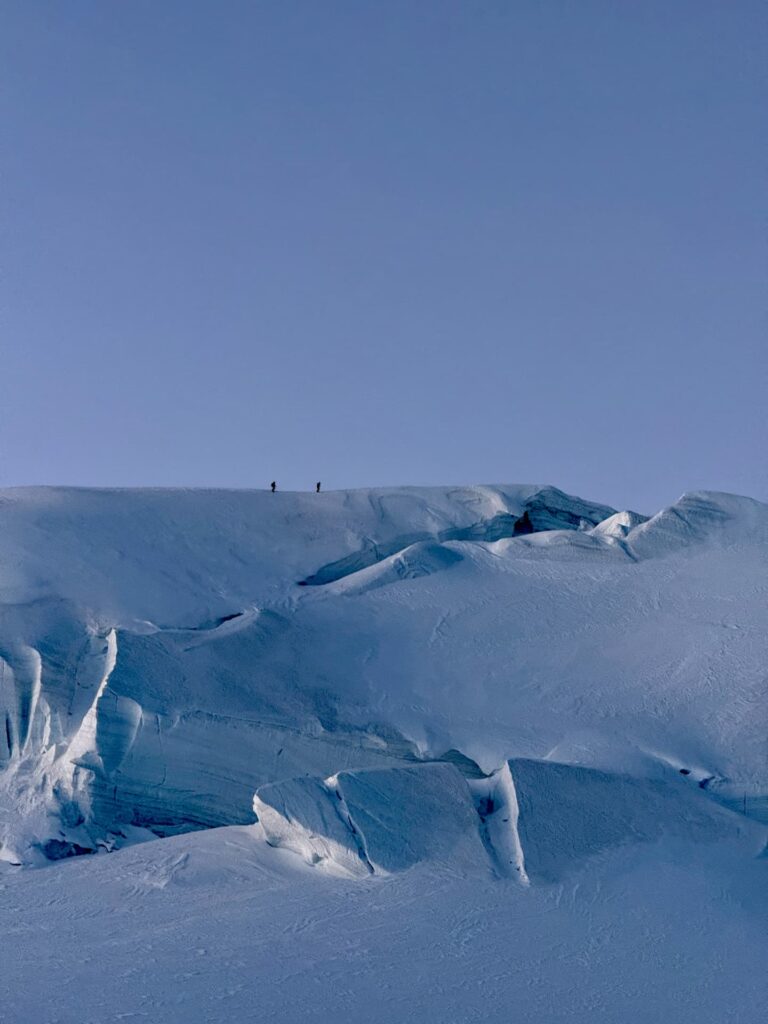 Zwei winzige Gestalten stehen auf einem weitläufigen, verschneiten Bergrücken unter einem strahlend blauen Himmel, während tiefe Gletscherspalten und strukturiertes Eis den Weg zum Gipfel hinaufführen – eine atemberaubende Szenerie, die an das Abenteuer der „Spaghetti Tour Monte Rosa“ erinnert.