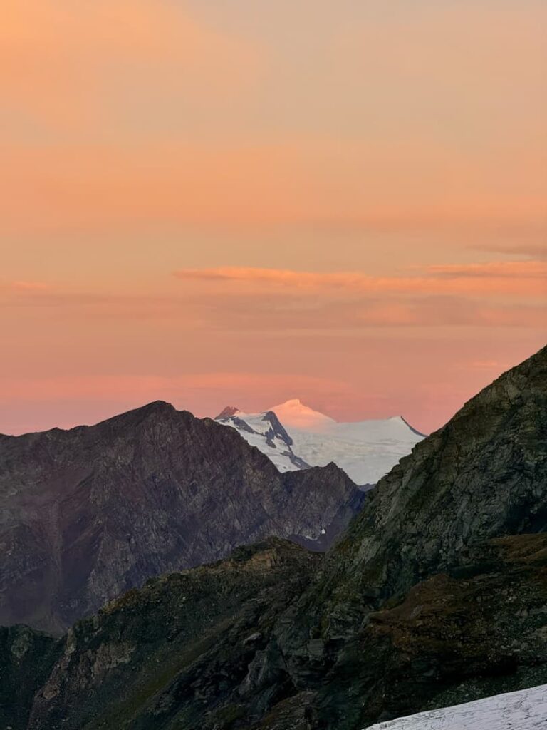 Ein schneebedeckter Berggipfel in der Ferne, umgeben von dunklen Felsketten, unter einem rosa-orangefarbenen Sonnenuntergangshimmel – eine unvergessliche Szene von einem „3000er Salzburger Land geführt“-Abenteuer.