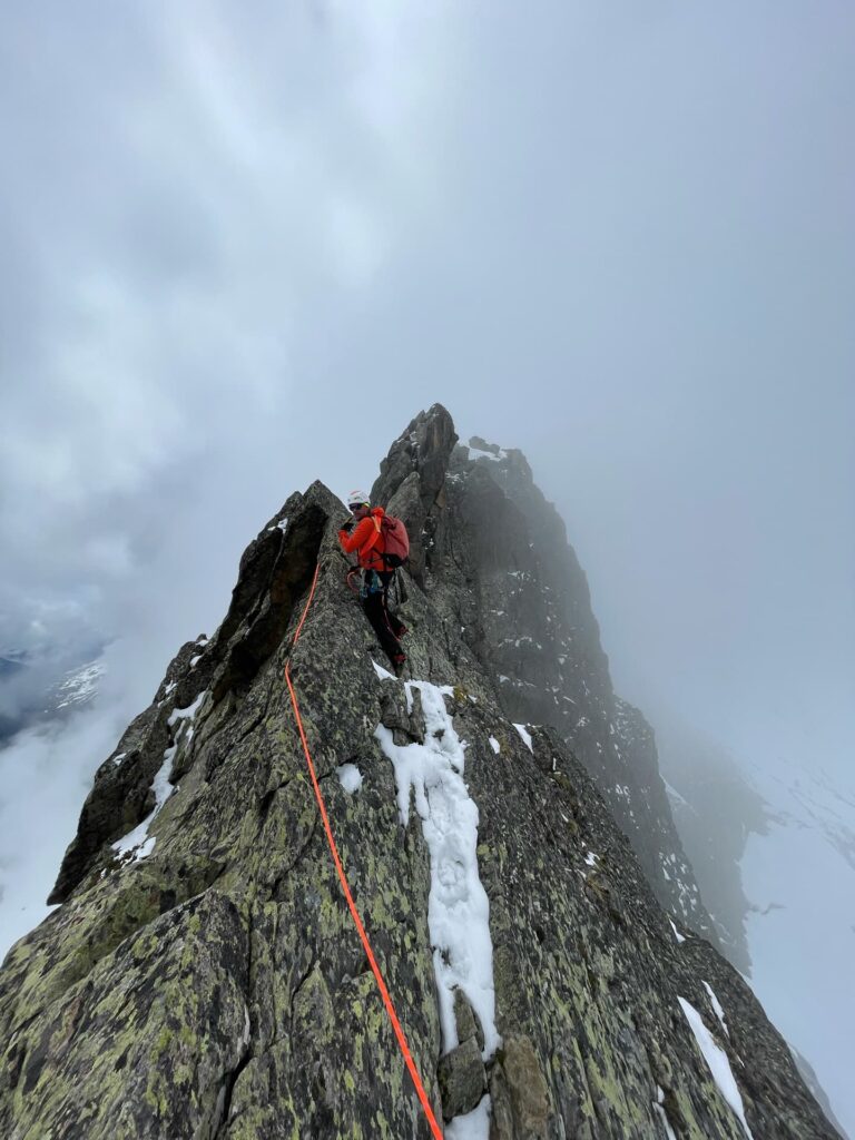 Ein Bergsteiger in einer roten Jacke erklimmt im Rahmen einer geführten Mont-Blanc-Expedition einen schmalen, felsigen und mit Schnee bedeckten Grat, gesichert durch ein orangefarbenes Seil, während im Hintergrund nebelverhangene Berge und Wolken zu sehen sind.