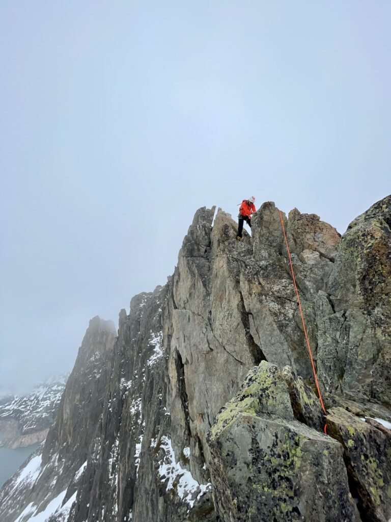 Ein Kletterer in einer orangefarbenen Jacke erklimmt mit einem Sicherungsseil einen steilen, felsigen Berggrat, umgeben von Nebel und Schneeflecken – ein Erlebnis, das an eine geführte Besteigung des Mont Blanc erinnert.