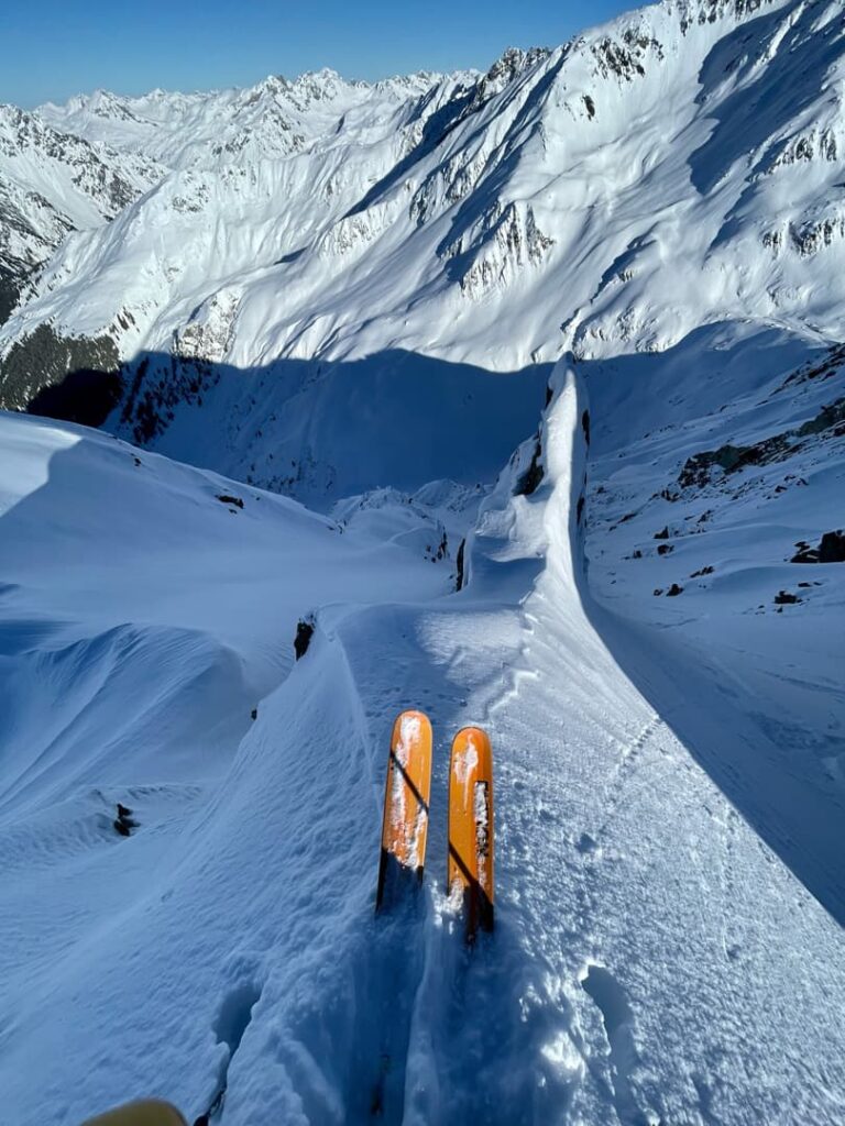 Der Blick eines Skifahrers auf leuchtend orangefarbene Skier, die auf einem schmalen, verschneiten Berggrat während eines Abenteuers der „Freeride Touren Alpen“ balancieren, während sich unter einem strahlend blauen Himmel steile Hänge und schroffe, schneebedeckte Gipfel bis in die Ferne erstrecken.