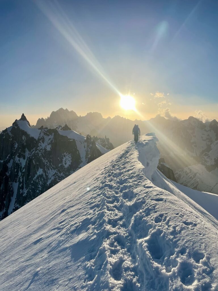 Eine Person wandert bei Sonnenaufgang oder Sonnenuntergang allein entlang eines verschneiten Berggrats, vor dem sich dramatische Gipfel und eine strahlende Sonne abzeichnen – eine Szene, die an ein geführtes Abenteuer am Mont Blanc erinnert.
