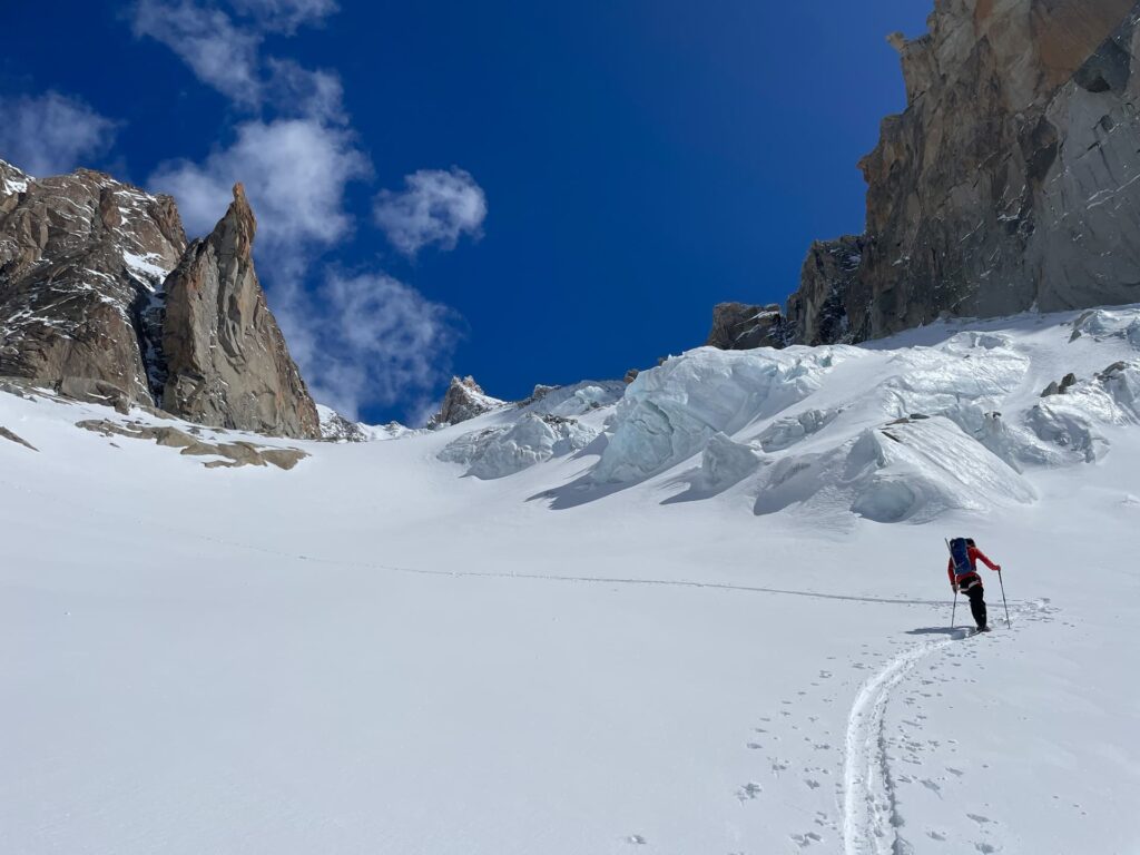 Ein einsamer Bergsteiger mit Rucksack wandert unter strahlend blauem Himmel mit vereinzelten Wolken einen verschneiten Hang hinauf, vorbei an zerklüfteten Felsen und vereisten Klippen, und verkörpert damit den Geist der geführten Skidurchquerungen in den Alpen.