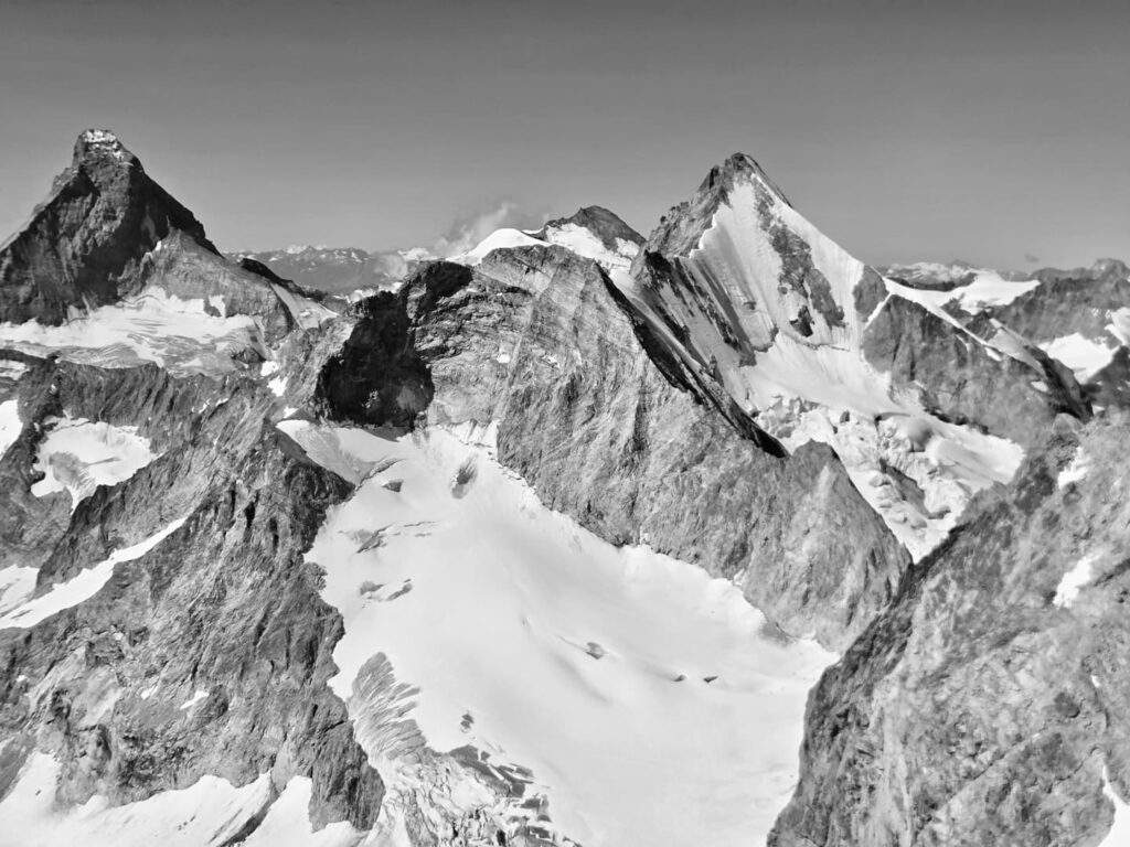 Schwarz-Weiß-Foto von schroffen, schneebedeckten Berggipfeln unter klarem Himmel, wobei schroffe Felswände und tiefe Schatten die dramatische Alpenlandschaft unterstreichen – eine Szene, die an eine Expedition zur Dom- und Dufourspitze erinnert.