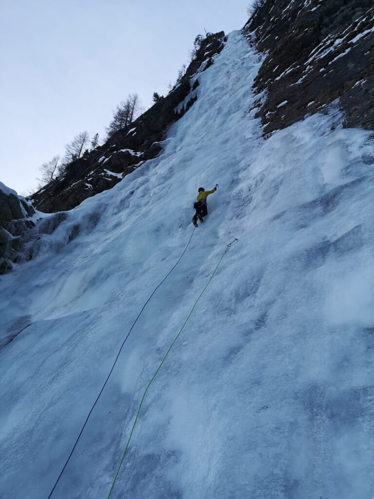 Ein Kletterer in einer gelben Jacke erklimmt mit Seilen und Eispickeln einen steilen, vereisten Hang, während er an einem gefrorenen Wasserfall Eisklettern betreibt.