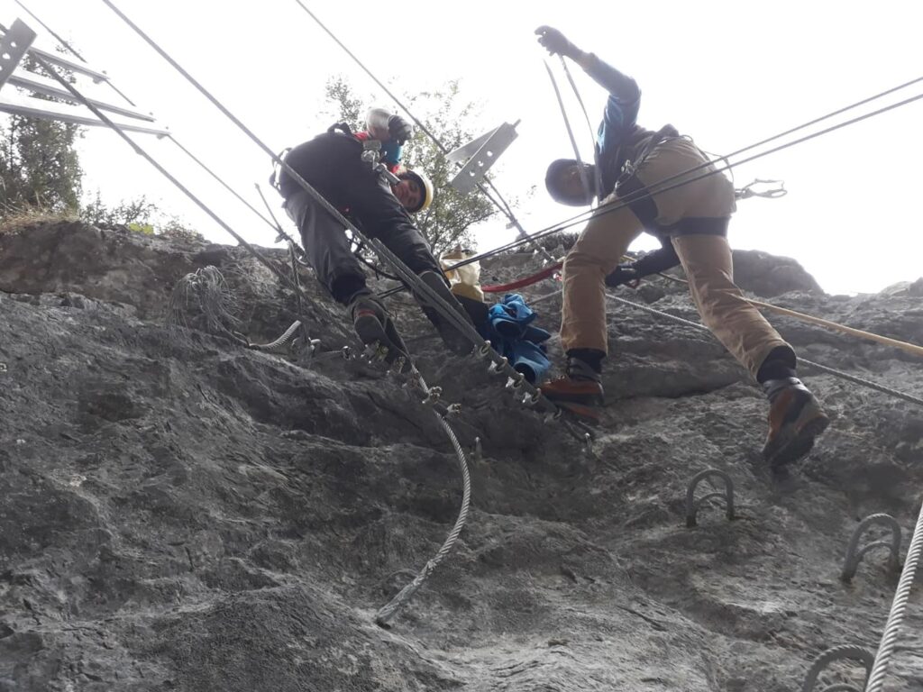 Zwei Personen in Kletterausrüstung erklimmen eine felsige Wand mithilfe von Seilen und am Fels befestigten Metallleitern – ein hervorragendes Beispiel für Seilarbeiten im alpinen Gelände. Das Foto wurde von unten aufgenommen und zeigt ihren Aufstieg sowie ihre Sicherheitsausrüstung.