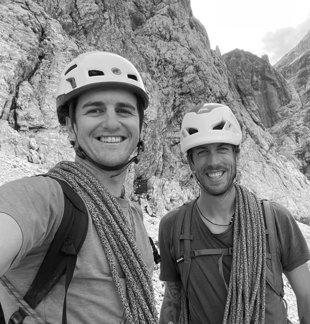 Zwei Männer mit Kletterhelmen und Klettergurten lächeln für ein Selfie in einer felsigen Berglandschaft. Das Foto ist in Schwarz-Weiß.