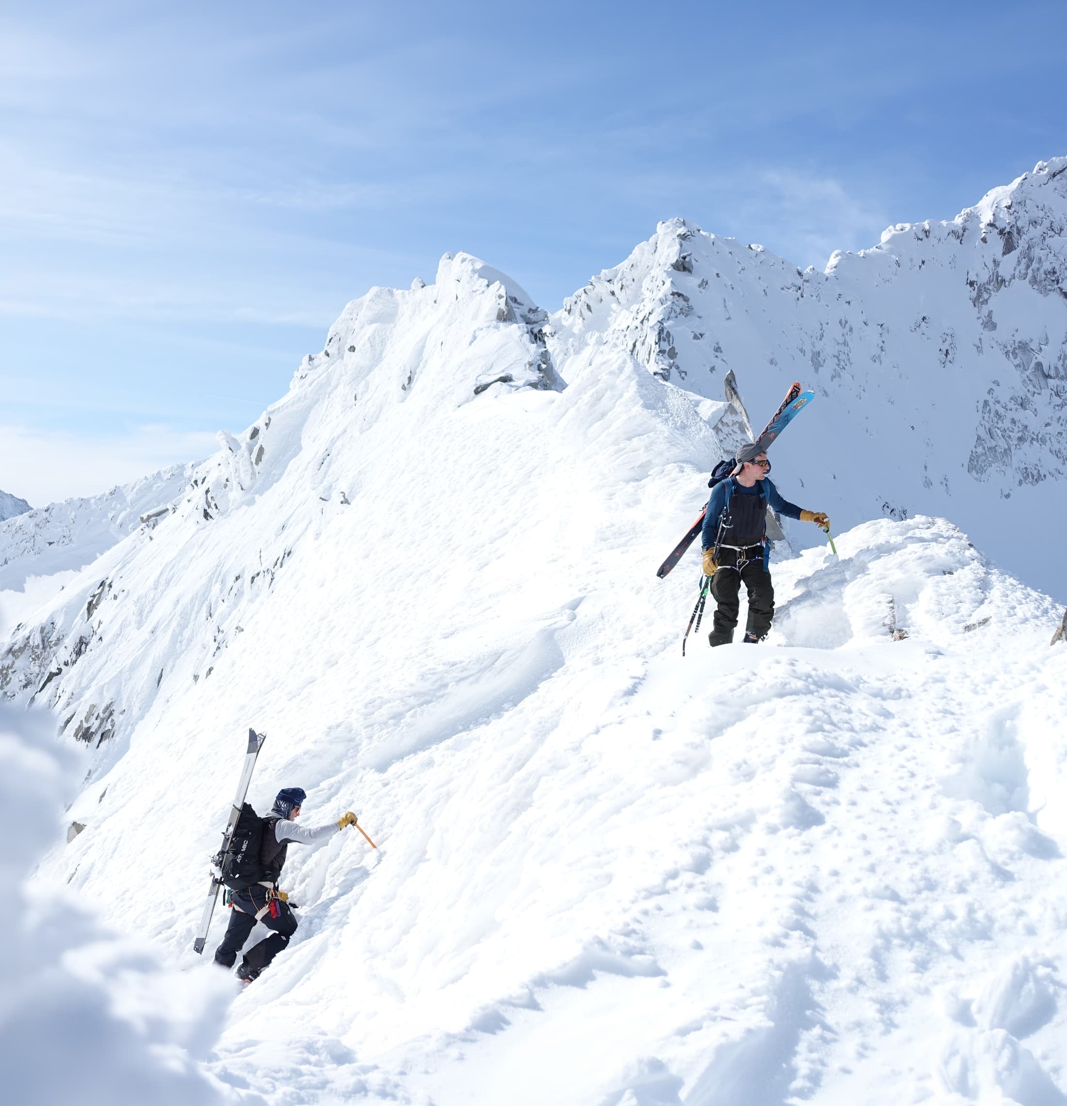 Zwei Menschen mit Skiern steigen unter strahlend blauem Himmel einen verschneiten Bergrücken hinauf, umgeben von hohen, schroffen, schneebedeckten Gipfeln – eine atemberaubende Szene, die an geführte Bergtouren durch unberührte Alpenlandschaften erinnert.