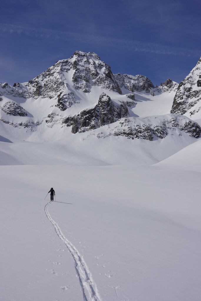 Ein einzelner Skifahrer gleitet unter strahlend blauem Himmel durch eine weite, schneebedeckte Landschaft auf schroffe, schneebedeckte Berge zu und hinterlässt dabei eine einzige Spur – was Gedanken an geführte Bergtouren weckt, die Abenteuer und atemberaubende Ausblicke auf die Alpen versprechen.