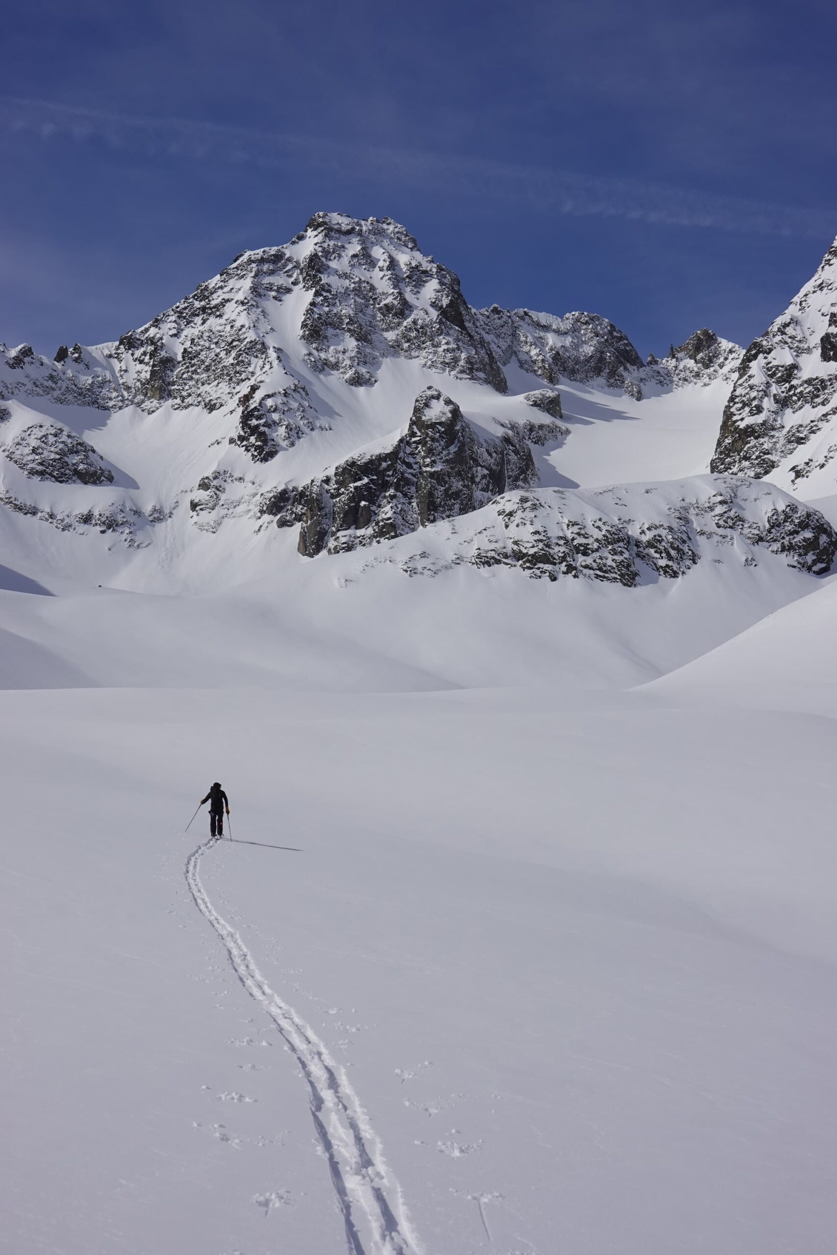 Ein einzelner Skifahrer gleitet unter strahlend blauem Himmel durch eine weite, schneebedeckte Landschaft auf schroffe, schneebedeckte Berge zu und hinterlässt dabei eine einzige Spur – was Gedanken an geführte Bergtouren weckt, die Abenteuer und atemberaubende Ausblicke auf die Alpen versprechen.